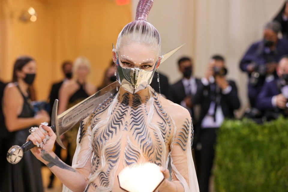 A person in an avant-garde dress and metal face mask holds a sword at a formal event, surrounded by photographers. Names are not available