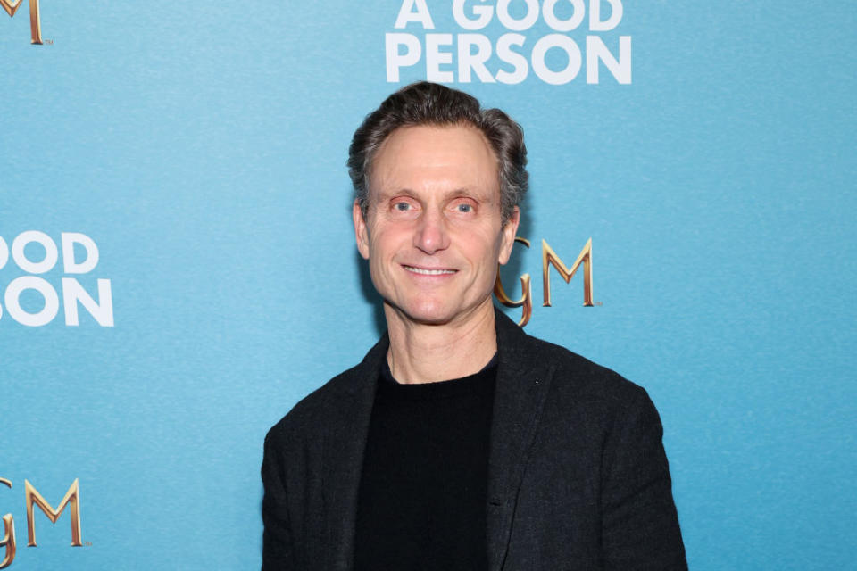 A person smiling in front of a backdrop reading "A Good Person." They are wearing a dark blazer over a black top