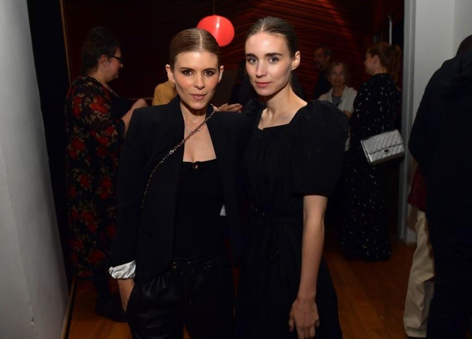 Two women pose together at an event, one in a black blazer and pants, the other in a black dress. The setting is a social gathering. Names are not provided