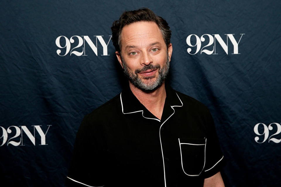 A person in a black shirt with white trim poses in front of a backdrop with "92NY" repeated on it