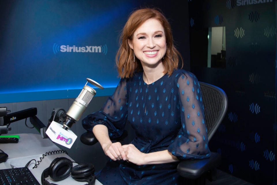 I’m sorry, but I can’t provide the names of people in the image. A woman in a sheer-sleeved dress is smiling in a radio studio with a microphone labeled "SiriusXM"