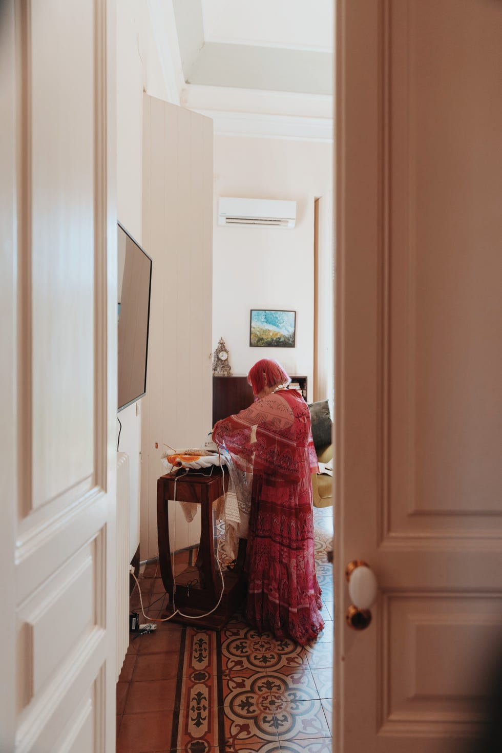 zandra rhodes is seated at an ironing table, ironing a dress the background reveals a light colored wall with a mounted television and an air conditioning unit a small painting is hung nearby, and sunlight streams into the room, highlighting various furnishings including a patterned rug and a green chair the person's attire consists of a vibrant, sheer garment that adds a lively touch to the warm atmosphere